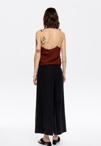 Bronzen satijnen camisole met bloemaccenten op de bandjes, gecombineerd met losse zwarte wijde broek. Gladde textuur, casual ontwerp.