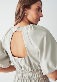 Robe gris clair avec des manches bouffantes, une découpe dans le dos et une taille froncée. Le tissu semble lisse avec une texture douce et un éclat subtil.