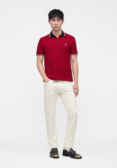 Jeune homme portant un polo rouge, un pantalon blanc et des chaussures noires avec des détails dorés, debout avec une main dans la poche sur fond blanc.