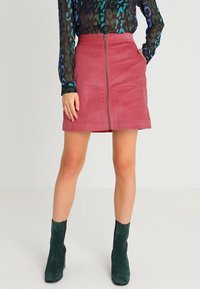 Paarse corduroy mini rok met een ritssluiting aan de voorkant, gecombineerd met een doorschijnende, gepatterniseerde top met lange mouwen en donkergroene enkellaarsjes.