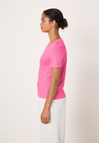 Giovane donna con maglietta rosa a maniche corte e pantaloni grigio chiaro in piedi di profilo contro uno sfondo semplice e chiaro.