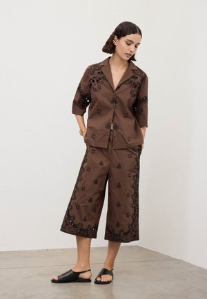 Frau trägt braune weite Hosen und ein passendes Hemd mit Knöpfen und schwarzer Stickerei, schwarze offene Sandalen, steht in einem neutralen Raum.