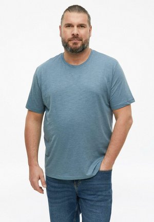 Man met baard en vastgebonden haar staat frontaal, draagt een blauw overhemd met korte mouwen en blauwe jeans, één hand in de zak.