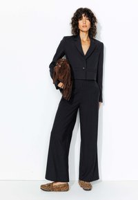 Blazer noir cintré sur un top en v, associé à un pantalon large à rayures, et des ballerines en imprimé léopard. Porte un sac en cuir frangé marron.