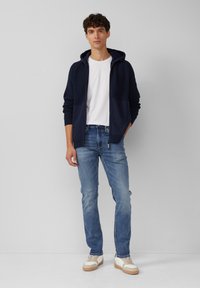Marinefarbener Zip-Hoodie mit geripptem Muster, weißes T-Shirt, hellblaue Jeans und beige Sneaker mit braunen Akzenten. Stehend vor neutralem Hintergrund.