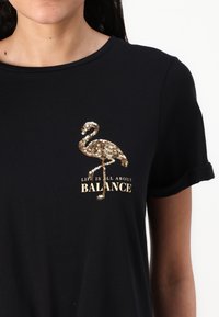 Black cotton t-shirt with rolled sleeves. Features a gold sequined flamingo design and text reading "LIFE IS ALL ABOUT BALANCE" in gold.
