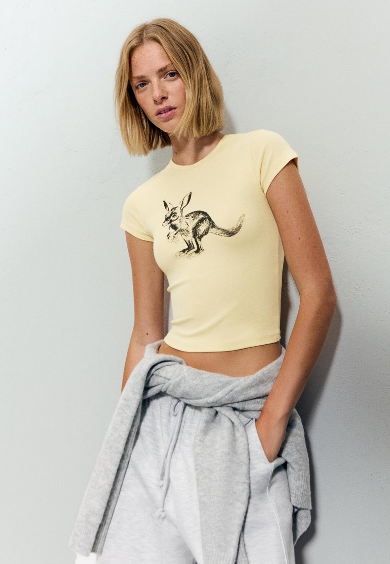 Žluté krátké tričko s krátkým rukávem a potiskem černého klokana, v kombinaci se šedými tepláky a šedým svetrem uvázaným kolem pasu.