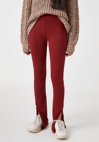 Pantaloni rossi aderenti con spacco laterale e una texture liscia, abbinati a un maglione lavorato a maglia spesso e multicolore e sneakers bianche.