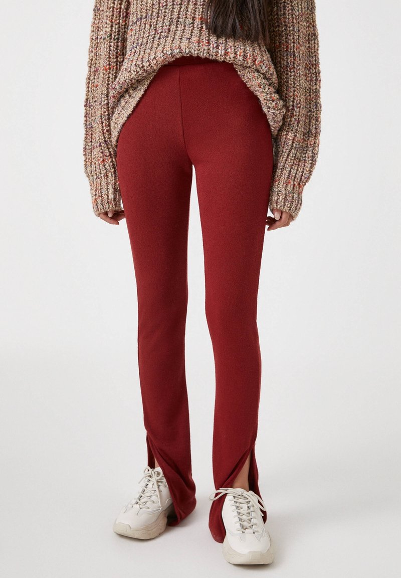 Pantaloni rossi aderenti con spacco laterale e una texture liscia, abbinati a un maglione lavorato a maglia spesso e multicolore e sneakers bianche.