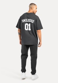 Camiseta de algodón negra con el texto blanco "SMILODOX 01" en la parte posterior, combinada con pantalones de chándal negros y zapatillas blancas, mostrando un ajuste relajado.