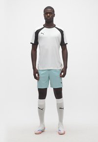 Άνδρας που στέκεται, φορώντας λευκό και μαύρο αθλητικό πουκάμισο Puma, ανοιχτό μπλε σορτς, λευκές κάλτσες μέχρι το γόνατο και λευκά ποδοσφαιρικά παπούτσια με μπλε και πορτοκαλί λεπτομέρειες.