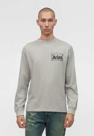 Junger Mann trägt ein hellgraues Langarmshirt mit dem Schriftzug "Widder" und einer Grafik auf der Brust, kombiniert mit blauen Jeans mit hellgrünen Mustern.