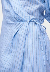 Φωτεινό γαλάζιο φόρεμα με λεπτές κάθετες ρίγες δεμένο στη μέση με κόμπο, που δείχνει την υφή και την πτώση του υφάσματος.