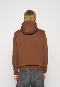Hnědá mikina s velkou kapucí, pružnými manžetami a lemem. Vyrobena z měkké tkaniny, má jednoduchý design a je kombinována s tmavými džíny.