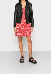 Černá kožená motorkářská bunda přes červené šaty s bílými květinovými vzory, doplněná o černé ploché sandály s přezkou.