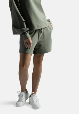 Henkilö, joka pukeutunut oliivinvihreään pitkähihaiseen paitaan ja vastaaviin shortseihin, seisoo taskut käsissä, jalassa valkoiset tennarit.