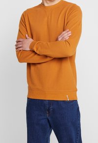 Sudadera de cuello redondo naranja con una tela texturizada, puños y dobladillo acanalados, y un pequeño detalle de logo cerca de la cintura. Combinada con jeans azules.
