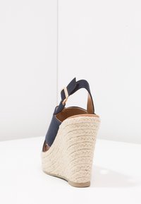 Marinblå kilklackssandal med en vävd espadrilleklack, ankelrem med spänne och ett slätt ovandelmaterial.