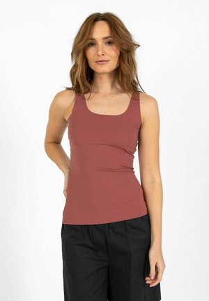 Vrouw met golvend bruin haar, dragend een mouwloos roestkleurig tanktop en zwarte losse broek, staand met één hand op haar heup.
