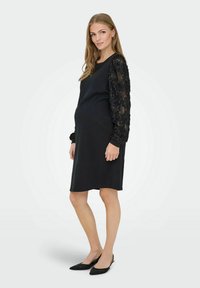 Robe de maternité noire avec de longues manches à sequins. La robe présente une coupe décontractée et un ourlet au niveau des genoux, associée à des ballerines noires à bout pointu.