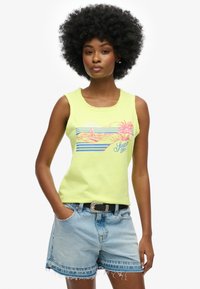 Lichtgele mouwloze top met een grafische print van palmbomen en golven, gecombineerd met denim shorts en een zwarte riem.