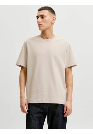 Rövid hajú férfi, aki bézs színű, túlméretezett pólót és sötét farmert visel, balra néz egyszerű, világos háttér előtt.