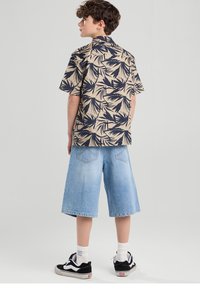 Camisa de manga corta beige con patrón de hojas en azul marino, combinada con pantalones cortos de mezclilla azul claro hasta la rodilla y zapatillas negras.