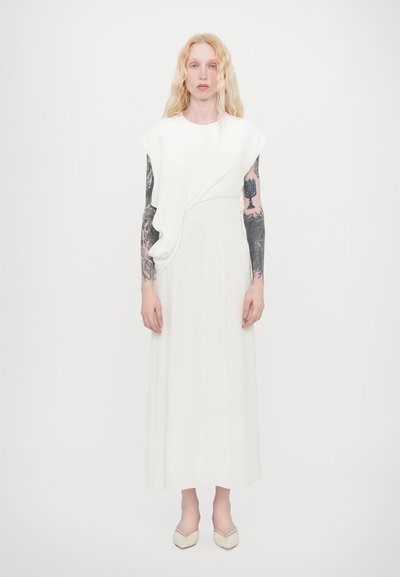Vestido blanco con drapeado asimétrico en el hombro, silueta larga y fluida, confeccionado en tejido suave, acompañado de zapatos planos beige.