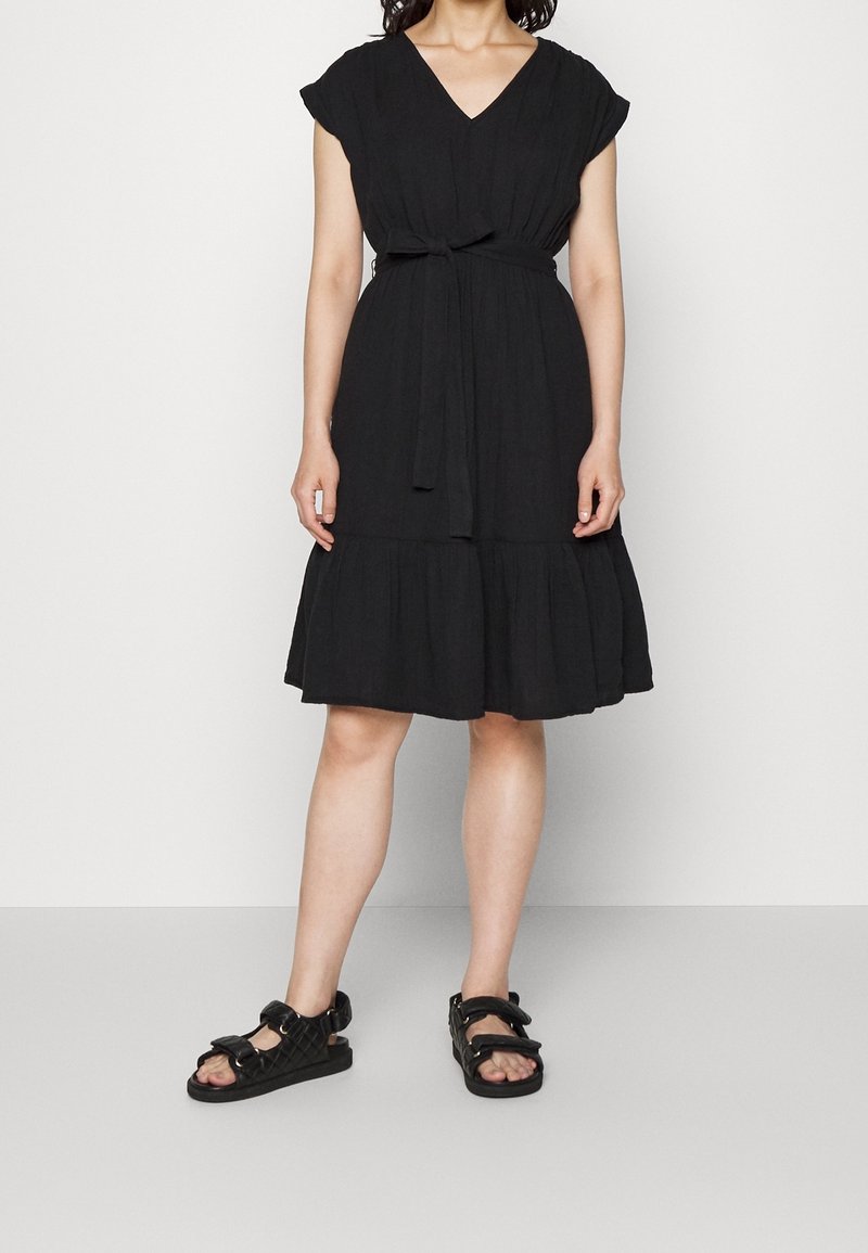 Femme portant une robe noire mi-longue avec une ceinture à nouer à la taille et des sandales noires à brides, debout devant un fond clair uni.