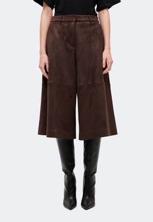 Femme portant un pantalon midi marron foncé taille haute et jambes larges, associé à des bottes noires montantes à bout pointu sur un fond uni.