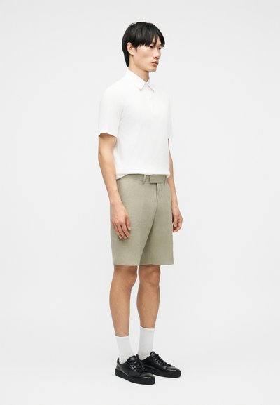 Άνδρας όρθιος, φοράει λευκό πόλο με κοντά μανίκια, ανοιχτό μπεζ σορτς μέχρι το γόνατο, λευκές κάλτσες και μαύρα αθλητικά παπούτσια, μπροστά σε απλό ανοιχτόχρωμο φόντο.