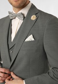Grauanzugjacke aus strukturiertem Stoff, begleitet von einer gepunkteten Fliege und einer floralen Reversnadel; ausgestattet mit einem weißen Einstecktuch mit grünen Akzenten.