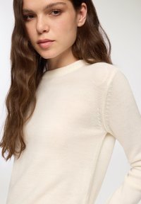 Sweter w kolorze kremowym, dziany, z prążkowanym okrągłym dekoltem, z długimi rękawami i fakturowanym materiałem. Szwy wzdłuż ramion.
