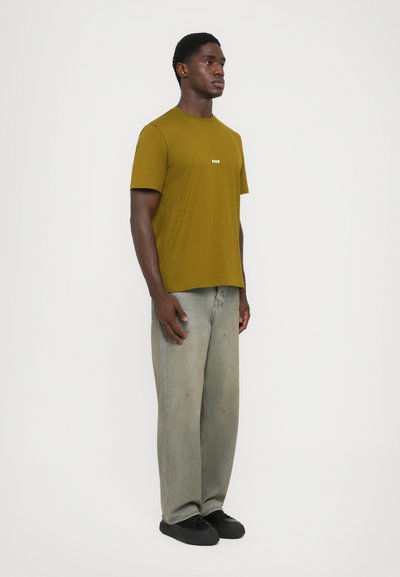 Olive green short-sleeve T-shirt with a small white logo, paired with light faded jeans and black sneakers. Model stands in profile.