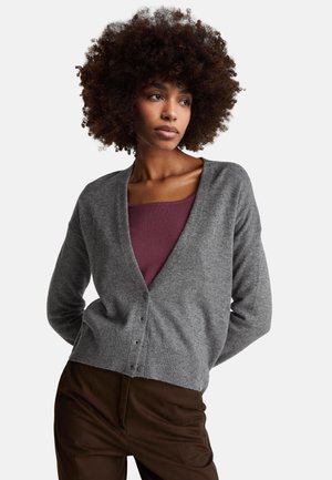 Frau mit lockigem Haar, die eine graue, geknöpfte Strickjacke über einem lila Oberteil und dunkelbraune Hosen trägt, vor einem schlichten weißen Hintergrund steht.