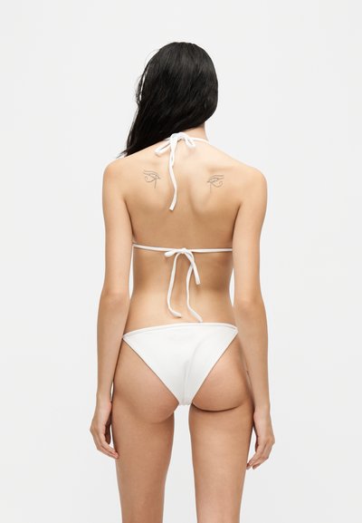 Femme aux longs cheveux foncés portant un bikini blanc vue de dos, montrant deux petits tatouages sur le haut du dos.