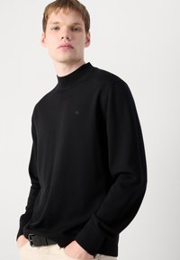 Črna pulover s stoječim ovratnikom, izdelan iz mehkega pletenega materiala. Ima majhen logotip na prsih ter rebraste manšete in rob.