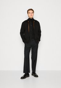 Black zip-up jacket with orange accents, worn over a black turtleneck. Paired with dark trousers and glossy black shoes.