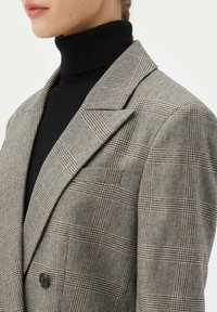 Grå hundtandsmönstrad kavaj med tvåknapprad, skarpa slag och en texturerad yta, lagd över en svart polotröja.