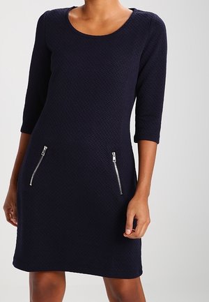 Vestido azul marino texturizado con mangas tres cuartos y bolsillos con cremallera expuesta a los lados. Escote redondo clásico y silueta ajustada.