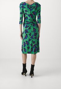 Grön klänning med blåa och röda fjärils mönster, trekvartsärmar, rynkad ryggdetalj och knälång skärning. Svarta ankelstövlar.