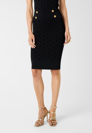 Femme portant une jupe noire mi-longue avec des boutons dorés et un tissu transparent à motifs, associée à des sandales à talons hauts dorées.
