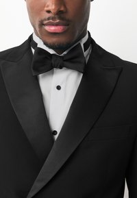 Black tuxedo jacket with satin lapels, white shirt, and black bow tie. Features black buttons and a functional pocket on the jacket.