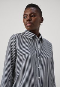 Švarkelis su kaklu ir ilgomis rankovėmis, pagamintas iš lengvos medžiagos su lygiu paviršiumi; juodai balti vertikalių dryžių raštai.