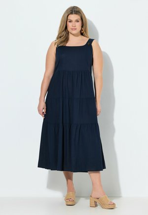 Frau trägt ärmelloses, marineblaues Stufen-Midikleid mit Knopfdetail an einem Träger und hellbraune, geflochtene Sandaletten mit Absatz, steht vor weißem Hintergrund.