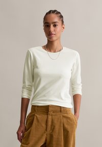 Frau mit geflochtenem Haar, die ein weißes Langarmshirt, eine silberne Kettenkette und braune Cordhosen trägt, mit den Händen in den Taschen.