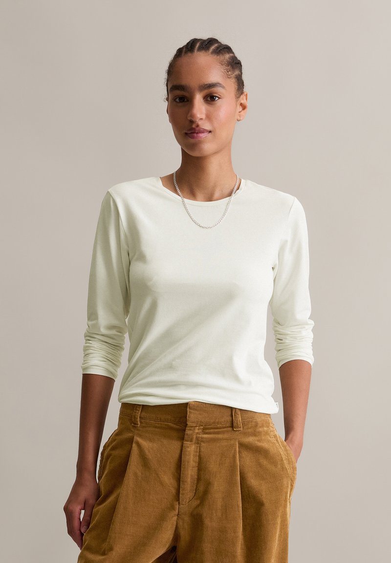 Frau mit geflochtenem Haar, die ein weißes Langarmshirt, eine silberne Kettenkette und braune Cordhosen trägt, mit den Händen in den Taschen.