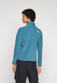 Blue fleece jacket with high collar, long sleeves, and a small logo on the back. Soft texture, mid-length, paired with black pants.
