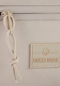 Világos bézs textúrájú táska cipzárral, csomózott zsinórral és egy téglalap alakú logószelvénnyel, amelyen az olvasható: "JOHNNY URBAN."