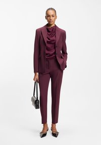 Burgundy tailored suit with a fitted blazer and matching trousers. Satin top, pointed black shoes, and a small black handbag. Smooth texture.